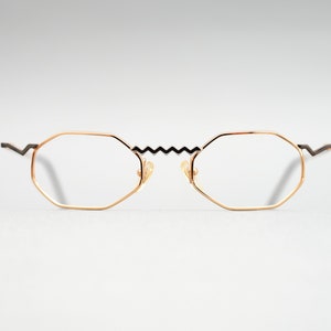 May include: Gold-tone octagon-shaped eyeglasses with black zig-zag bridge and black arms.