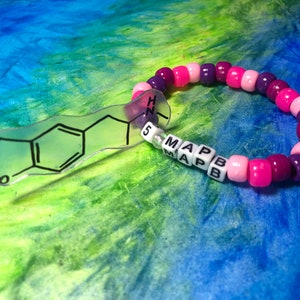 May include: A beaded bracelet with a clear charm featuring a black outline of a chemical structure. The bracelet has pink, purple, and white beads. The charm has the letters "5 MAPB" on it.