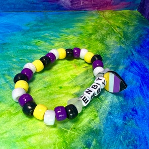 May include: A beaded bracelet featuring a repeating pattern of black, yellow, white, and purple beads. The bracelet has a charm with the non-binary flag and the letters "ENBY" on white letter beads. The background is a colorful blend of blue, green, and purple.