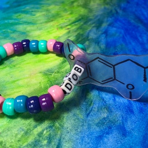 May include: A beaded bracelet with a clear, blue charm displaying a chemical structure and the letters "DOB". The bracelet features beads in pink, purple, teal, and blue. The charm is a light blue colour with black lines.