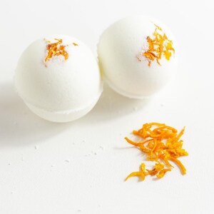 May include: Two white bath bombs with orange zest on a white background.