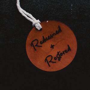 May include: A brown leather tag with white string attached. The tag has the words "Redeemed + Restored" written in black ink.