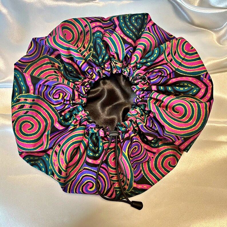 May include: A black satin lined sleep cap with a pink, green, and gold swirl pattern. The cap has a drawstring closure.