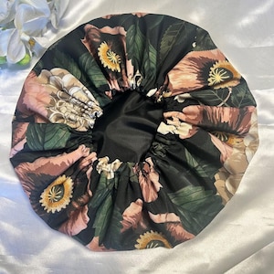 Op de afbeelding: Een zwarte satijnen bonnet met een bloemenprint en een elastische rand. Het patroon bevat grote bloemen in roze, perzik, geel en groen. De binnenkant van de bonnet is effen zwart.