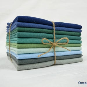 Op de afbeelding: Een stapel gevouwen stoffen vierkanten in tinten blauw, groen en grijs, vastgebonden met touw. De stoffen vari&euml;ren van diep marineblauw tot lichtblauw en grijs, waardoor een op de oceaan ge&iuml;nspireerd palet ontstaat. De tekst "Ocean Mix" is zichtbaar.