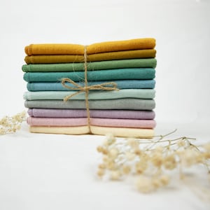 Op de afbeelding: Een stapel kleurrijke, rechthoekige vilt vierkanten vastgebonden met touw. De vilt vierkanten zijn in tinten geel, groen, blauwgroen, blauw, grijs, lavendel en roze. De stapel staat op een witte achtergrond.