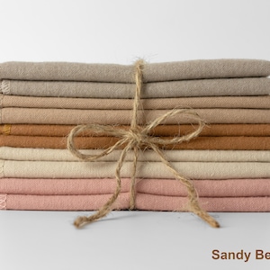 Op de afbeelding: Een stapel gevouwen stoffen in verschillende neutrale kleuren, waaronder beige, bruin en roze, samengebonden met touw. De stof is netjes gerangschikt en de algehele esthetiek is eenvoudig en elegant. De tekst "Sandy Beach" staat onderaan.