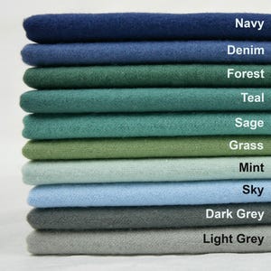 Op de afbeelding: Een stapel stoffen stalen in verschillende kleuren, waaronder marineblauw, denim, bosgroen, blauwgroen, salie, gras, mint, hemelsblauw, donkergrijs en lichtgrijs. Elk staal is gelabeld met de bijbehorende kleurnaam.