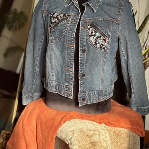 May include: A blue denim jacket with a cropped fit and embellished pockets. The pockets are decorated with rhinestones and sequins.