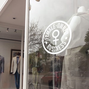 May include: Exterior shot of a storefront with mannequins displaying suits. A large white circular sign on the window reads "FEMALE-OWNED BUSINESS" with a female symbol. The building has a brick facade and a blue awning.