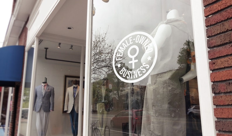 May include: Exterior shot of a storefront with mannequins displaying suits. A large white circular sign on the window reads "FEMALE-OWNED BUSINESS" with a female symbol. The building has a brick facade and a blue awning.