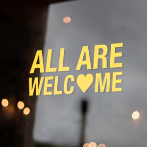 May include: A yellow sign on a glass door with the text "ALL ARE WELCOME" and a yellow heart.