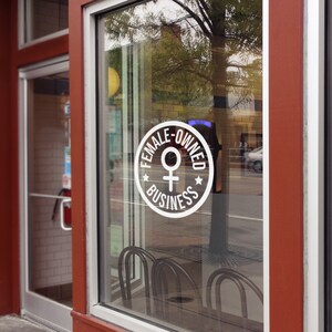May include: Exterior shot of a business storefront with a large window. A white circular sign on the glass reads "FEMALE-OWNED BUSINESS" with a Venus symbol. The building's trim is a deep red, and the glass reflects the street scene.