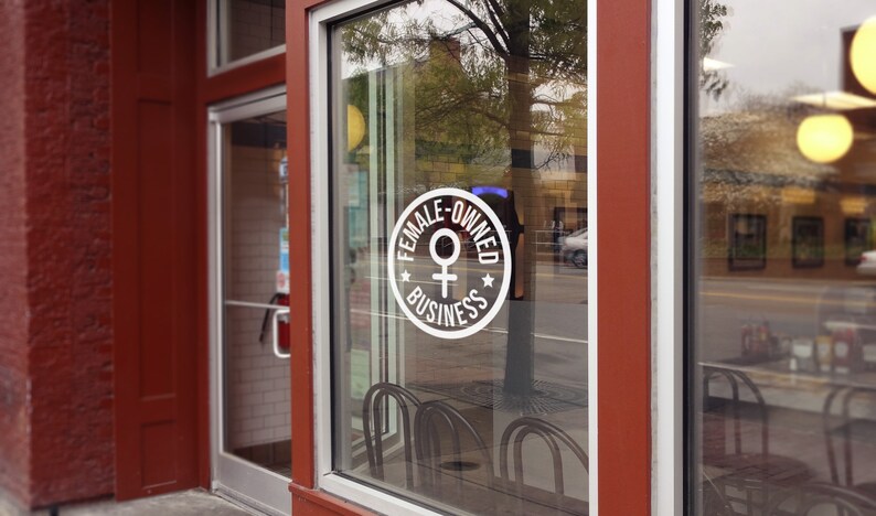 May include: Exterior shot of a business storefront with a large window. A white circular sign on the glass reads "FEMALE-OWNED BUSINESS" with a Venus symbol. The building's trim is a deep red, and the glass reflects the street scene.