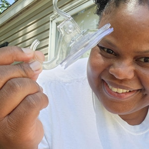 May include: Clear plastic suction cup hook with a curved design. The hook is held up by a person wearing a white t-shirt with the text "for they shall" printed on it.