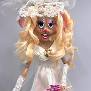 May include: A handmade doll with a mouse face, wearing a white lace dress, a white lace hat with a feather, and white lace gloves. The doll is holding a pink fan.