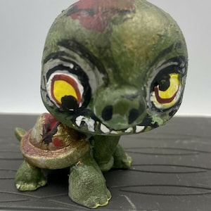 May include: A green and gold painted turtle figurine with a zombie-like face. The turtle has red and yellow eyes, a large toothy grin, and a green shell with gold accents.