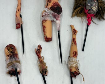 Puede incluir: Colección de espeluznantes decoraciones de Halloween con partes del cuerpo desmembradas. El conjunto incluye un torso, una cabeza, brazos y piernas, todos unidos a palos negros. Las partes del cuerpo están pintadas con sangre y envueltas en gasa y cuerda.