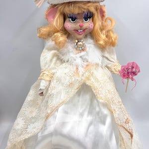 May include: A white doll with blonde hair and a pink nose wearing a white dress with lace and a white hat with white flowers. The doll is holding a pink fan.