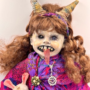 May include: A handcrafted doll with a grotesque, aged face, adorned with horns and a purple and pink patterned scarf. The doll has red hair, a long tongue, and a pentagram pendant. A small, stitched bunny toy is clutched nearby. The doll's design evokes a gothic or fantasy theme.