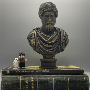 Puede incluir: Un busto de bronce de Marco Aurelio sobre un pedestal, con un libro titulado "Marcus Aurelius Meditations" y un libro titulado "Fowler's Archeology" debajo. Un peque&ntilde;o b&uacute;ho blanco y una figura de Lego con una espada est&aacute;n frente al busto.