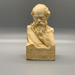 May include: A beige bust sculpture of a man with a beard and a serious expression. The sculpture is made of a smooth, textured material and is set on a square base.