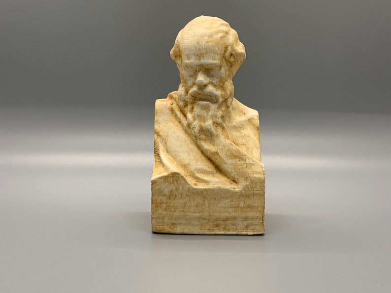 May include: A beige bust sculpture of a man with a beard and a serious expression. The sculpture is made of a smooth, textured material and is set on a square base.