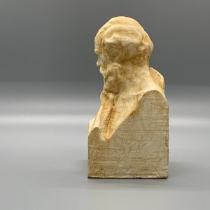 May include: A white plaster bust of a man with a beard and a textured surface. The bust is on a square base.