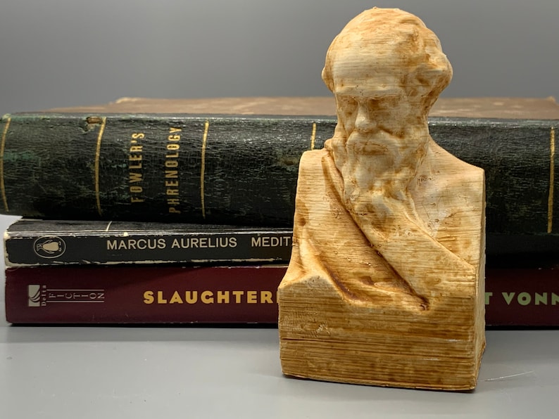 May include: A wooden bust of a man with a beard and a long, flowing robe. The bust is sitting on a stack of books. The books have titles that include "Fowler's Phrenology", "Marcus Aurelius Meditations", and "Slaughterhouse Five".