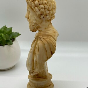 May include: A cream-colored bust sculpture of a man with curly hair and a beard. The bust is on a round base and is made of a smooth material.