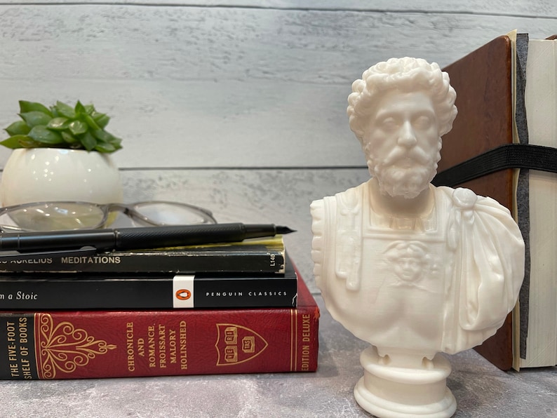 May include: White plaster bust of a man with a beard and short hair. The bust is on a white pedestal and is sitting on a stack of books. The books are titled "Meditations", "A Stoic", "Chronicle and Romance", and "Froissart Malory Holinshed".