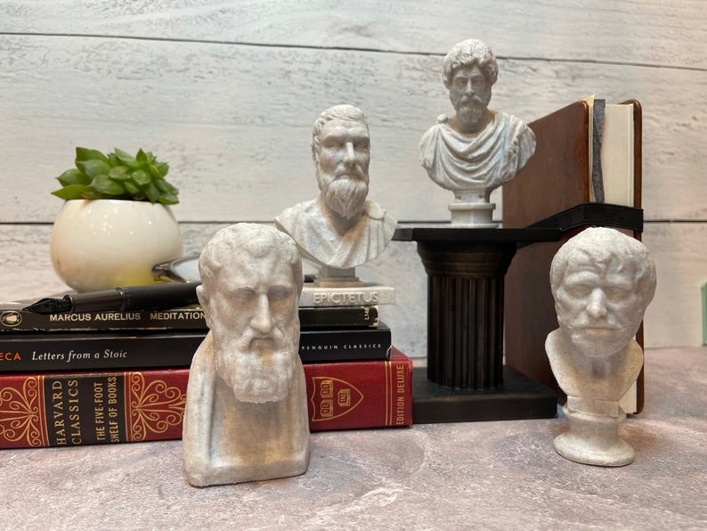 Puede incluir: Cinco bustos de yeso blanco de fil&oacute;sofos famosos, entre ellos Epicteto, Marco Aurelio y S&eacute;neca, se exhiben en una estanter&iacute;a con libros sobre estoicismo. Los bustes son todos de estilo cl&aacute;sico, con rasgos realistas y t&uacute;nicas fluidas.