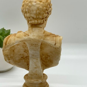 May include: A cream-colored bust sculpture of a person with short, curly hair. The bust is made of a smooth, textured material and is mounted on a round base.