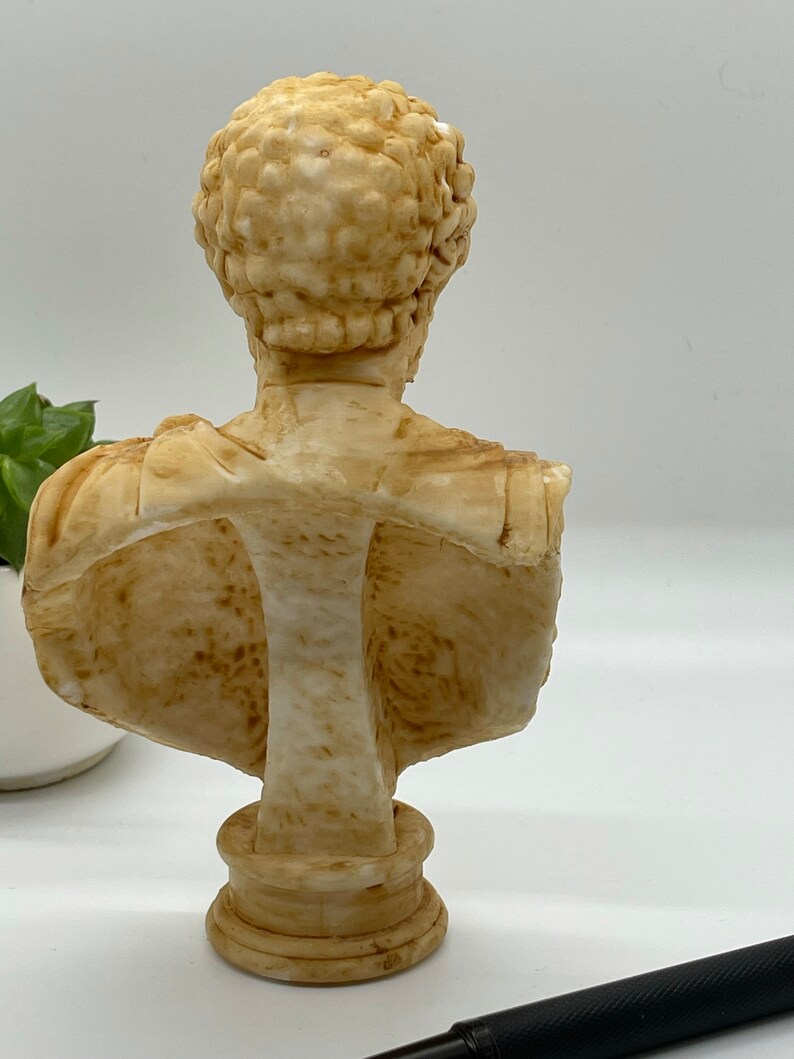 May include: A cream-colored bust sculpture of a person with short, curly hair. The bust is made of a smooth, textured material and is mounted on a round base.