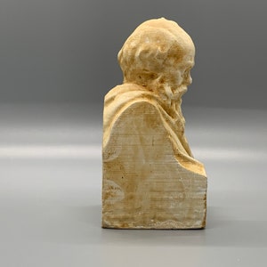 May include: A beige plaster bust of a bearded man with a detailed face and hair. The bust is on a rectangular base.