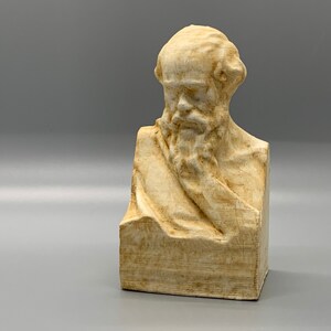 May include: A beige bust sculpture of a man with a beard and a draped cloth. The sculpture is made of a textured material and has a slightly weathered appearance.