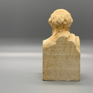 May include: A beige plaster bust of a person with a short, curly hairstyle. The bust is on a rectangular base.