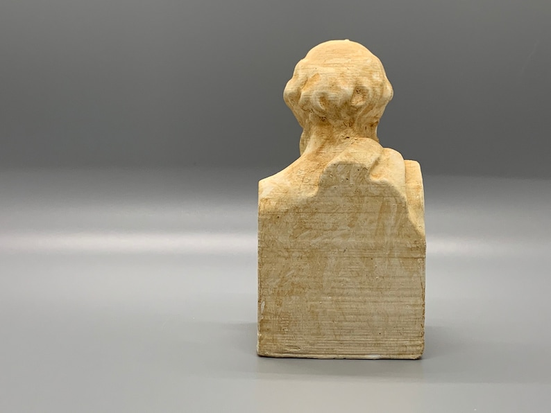 May include: A beige plaster bust of a person with a short, curly hairstyle. The bust is on a rectangular base.