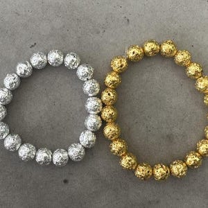 Könnte beinhalten: Vier elastische Armbänder mit kugelförmigen Perlen. Das erste ist weiß mit goldenen Buchstabenanhängern C, B und T. Das zweite hat metallisch silberne Perlen. Das dritte hat metallisch goldene Perlen. Das vierte ist schwarz mit einem silbernen Buchstaben T.