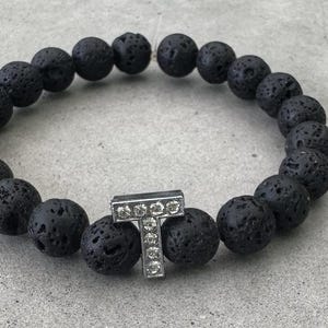May include: A circular stretch bracelet composed of black, porous lava stone beads. The bracelet features a single silver-toned metal charm in the shape of the capital letter T, which is embedded with small, clear, sparkling rhinestones.