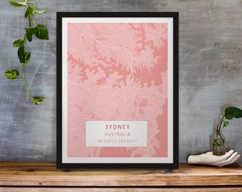 May include: A framed map of Sydney, Australia, with a pink background and white lines showing the city streets. The map is framed in black and has a white label with the text "SYDNEY AUSTRALIA 33.920'S / 151.157'E".
