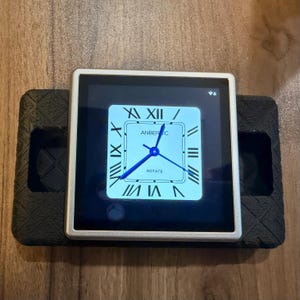 May include: A silver square smartwatch with a black screen showing a clock face with Roman numerals and the text "ANBERNIC" and "ROTATE". The watch is on a black textured base.