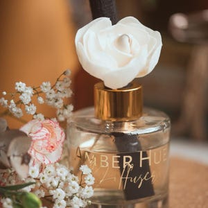 May include: A clear glass diffuser bottle with a gold cap and a white flower. The bottle has a black label with "AMBER HUE" and "Diffuser" in gold. White and pink flowers are arranged around the bottle. The bottle is filled with liquid.