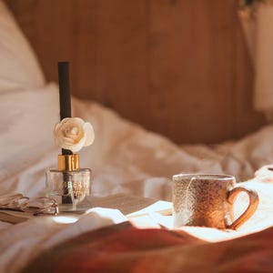 Könnte beinhalten: Eine transparente Glas-Diffusorflasche mit einer weißen Blume und einem schwarzen Schilfrohr, neben einer strukturierten braunen Tasse mit einem gebogenen Griff. Die Szene spielt sich auf einem Bett mit einem Buch und einer Brille ab.