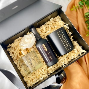 Könnte beinhalten: Eine schwarze Geschenkbox mit einem goldenen Band und dem aufgedruckten Text "Amber Hue". Die Box enthält eine klare Glasflasche mit goldenem Deckel, eine schwarze Flasche mit goldenem Deckel und eine schwarze Kerze mit goldenem Deckel. Die Kerze trägt den aufgedruckten Text "Amber Hue".