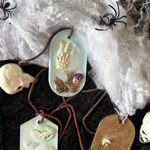 Puede incluir: Colgantes temáticos de Halloween con diseños espeluznantes. Los colgantes presentan una mano de esqueleto, murciélagos y fantasmas, sobre un fondo de telarañas blancas y redes negras. Calaveras decorativas y arañas de plástico se suman a la estética espeluznante.