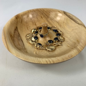 May include: A wooden ring dish with a natural wood grain finish. The dish has a small centre post for holding rings. A gold bracelet with black stones and charms is resting on the dish.