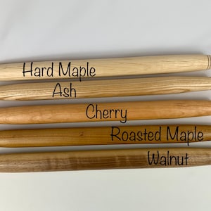 May include: Five wooden rolling pins, each made from a different type of wood: hard maple, ash, cherry, roasted maple, and walnut. The rolling pins are arranged vertically, with the hard maple rolling pin at the top and the walnut rolling pin at the bottom.