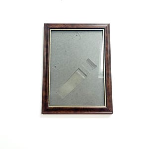 May include: A rectangular picture frame with a dark brown wooden frame and a gold-colored inner edge. The frame has a gray backing and is empty. The frame is hanging on a white wall.