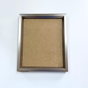 Puede incluir: Un marco de fotos plateado con un respaldo de cartón marrón.